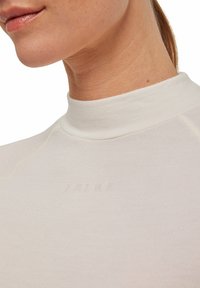 Top bianco a maniche lunghe con collo alto, realizzato in tessuto testurizzato e con un logo stampato discreto. Primo piano dei dettagli del colletto e della cucitura della spalla.