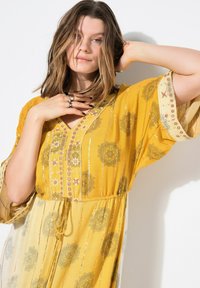 Kaftano giallo con motivi floreali, texture plissettata e scollatura a V. Presenta bottoni decorativi e una vita elastica per una migliore vestibilità.