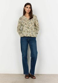 Blomstret langærmet top i creme med grønne og blå mønstre, parret med blå straight-leg jeans og brune åbne sko.