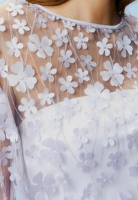 Transparenter lila Netzstoff mit bestickten floralen Applikationen, Rundhalsausschnitt und glattem weißen Stoff darunter.