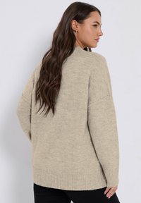 Maglione oversize beige caratterizzato da una trama morbida, polsini e orlo a coste, e un collo alto. La vista posteriore mostra una vestibilità ampia e rilassata.