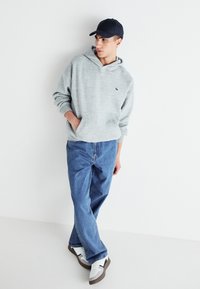 Sudadera gris con bolsillo frontal, superpuesta sobre jeans de color azul claro. Zapatillas blancas con detalles grises. Gorra negra completa el atuendo casual.