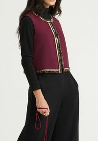 Veste sans manches bordeaux avec ornements perlés, associée à un haut noir à manches longues. Tissu lisse avec une forme structurée et devant ouvert.
