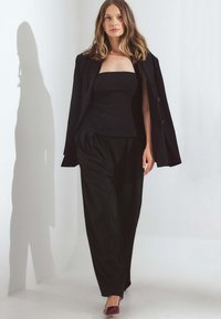 Czarna dopasowana marynarka na czarnym przylegającym topie i luźnych spodniach. Głębokie burgundowe obcasy. Gładki materiał, wyraźna sylwetka i czyste linie.