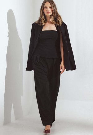 Schwarze maßgeschneiderte Blazer über einem schwarzen, figurbetonten Oberteil und lockeren Hosen. Dunkelburgunderfarbene Absatzschuhe. Glatter Stoff, strukturierte Silhouette und klare Linien.