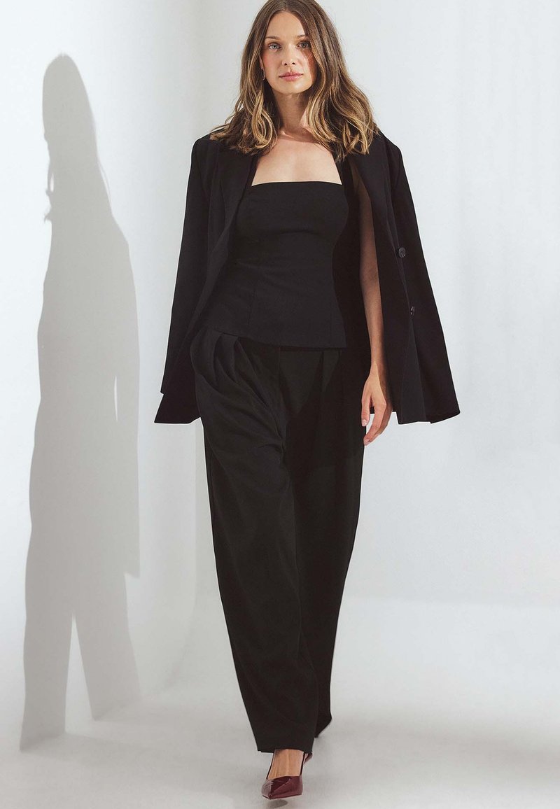 Czarna dopasowana marynarka na czarnym przylegającym topie i luźnych spodniach. Głębokie burgundowe obcasy. Gładki materiał, wyraźna sylwetka i czyste linie.