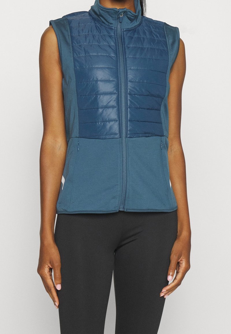 Ärmlös blå väst med quiltad front, dragkedjestängning, sidofickor och hög krage. Tillverkad av en blandning av slät och texturerad tyg.