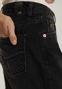 Czarne dżinsy o slim fit, z tylną kieszenią i subtelną metką marki. Tekstura jest lekko zużyta, co podkreśla ich swobodny wygląd.