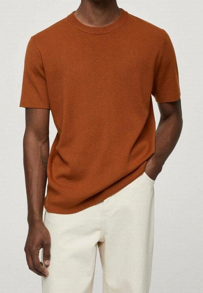 Homme portant une chemise en maille à manches courtes couleur rouille avec un pantalon écru, une main dans la poche, debout devant un fond neutre.