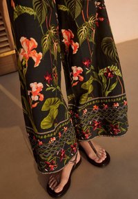 Pantalon large noir à motif floral vert et rouge, porté avec des sandales marron foncé à brides entre les orteils sur un sol clair.