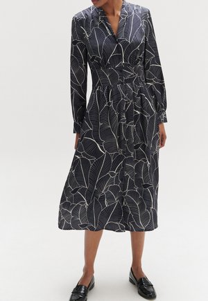 Marineblaues Kleid mit weißen Blättern, langen Ärmeln, kragenförmigem Ausschnitt und taillierter Taille, das bis zur Wadenmitte reicht. Sichtbare schwarze Loafers.