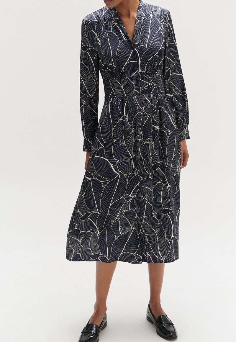 Marineblaues Kleid mit weißen Blättern, langen Ärmeln, kragenförmigem Ausschnitt und taillierter Taille, das bis zur Wadenmitte reicht. Sichtbare schwarze Loafers.