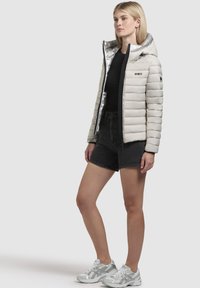 Lichtgewicht pufferjack in gebroken wit met een glanzende zilveren voering, voorzien van een capuchon en ritsluiting. Draag het met zwarte high-waisted shorts en zilveren sneakers.