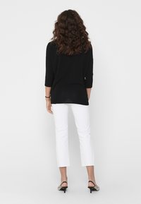 Schwarzes Strickoberteil mit Dreiviertelärmeln, kombiniert mit weißen, kurzen Hosen mit rohen Säumen. Das Model trägt schwarze und graue, gemusterte Absatzschuhe.