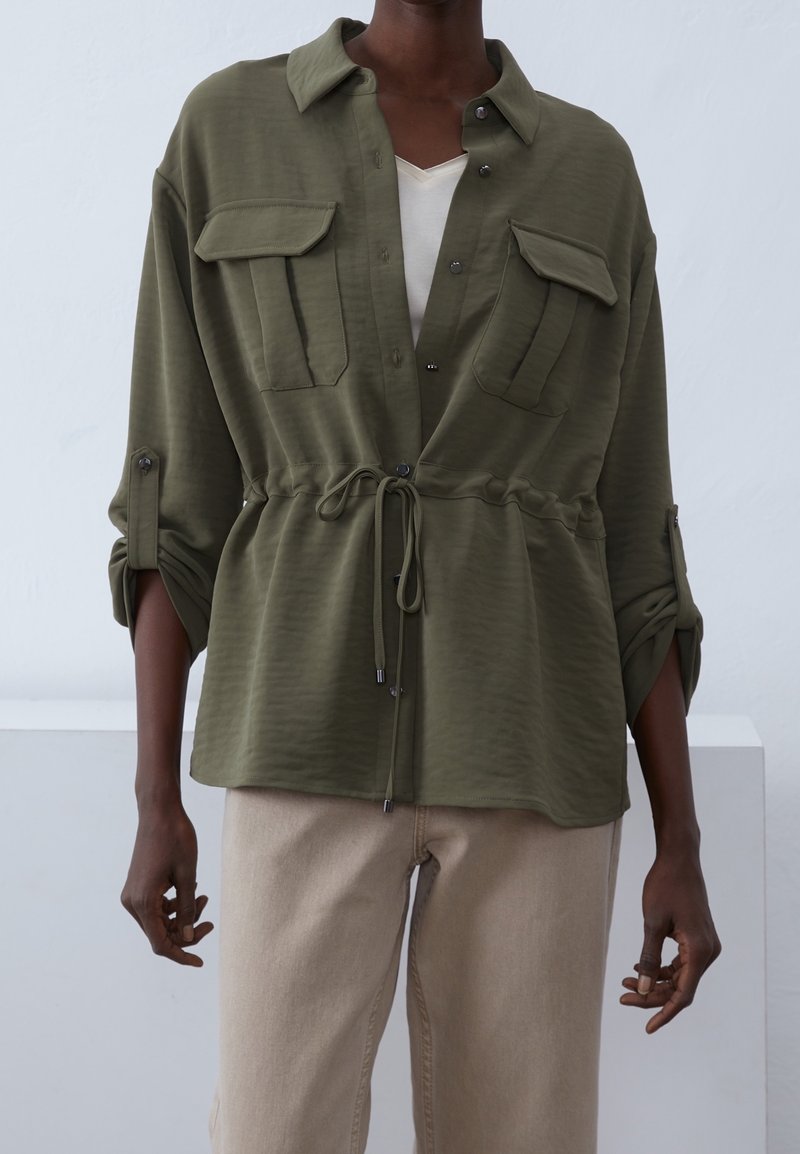 Veste-chemise légère vert olive avec manches retroussées, deux poches poitrine, taille à cordon de serrage, et texture subtile. Sous-vêtement neutre visible.