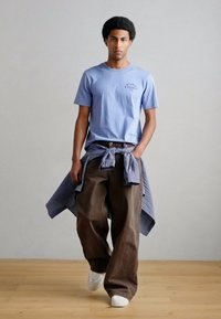 Ανοιχτό μπλε μπλουζάκι, καφέ παντελόνια με φαρδύ πόδι και δεμένο καρό πουκάμισο. Λευκά αθλητικά παπούτσια, απαλή υφή, μινιμαλιστικός σχεδιασμός, στέκεται πάνω σε ξύλινο πάτωμα.