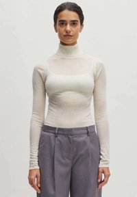 Weißes, langärmeliges Turtleneck-Oberteil aus transparentem Stoff; tailliert, mit runder Ausschnitt. Kombiniert mit grauen, hochgeschnittenen Hosen mit Falten.