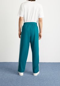 Tealfärgade mjukisbyxor med vita sidosträck, elastisk midja och lös passform, i kombination med en enkel vit t-shirt, sedd bakifrån.