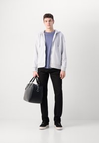 Sweatshirt zip cinza claro, t-shirt azul, calças pretas, ténis pretos, segurando uma mala de couro preta com duas alças, em pé sobre um fundo branco.