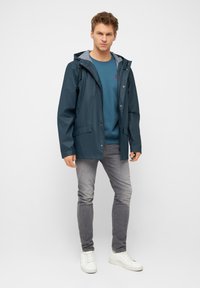 Giacca impermeabile blu navy con cappuccio regolabile, foderata in grigio e dotata di due tasche frontali. Abbinata a un maglione blu, jeans grigi e sneaker bianche.