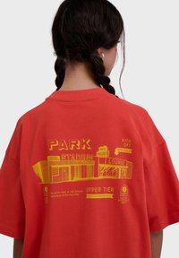 Persona con cabello trenzado que lleva una camiseta roja con un gráfico amarillo de un estadio y texto que incluye "PARK", "STADIUM" y "LOUNGE".