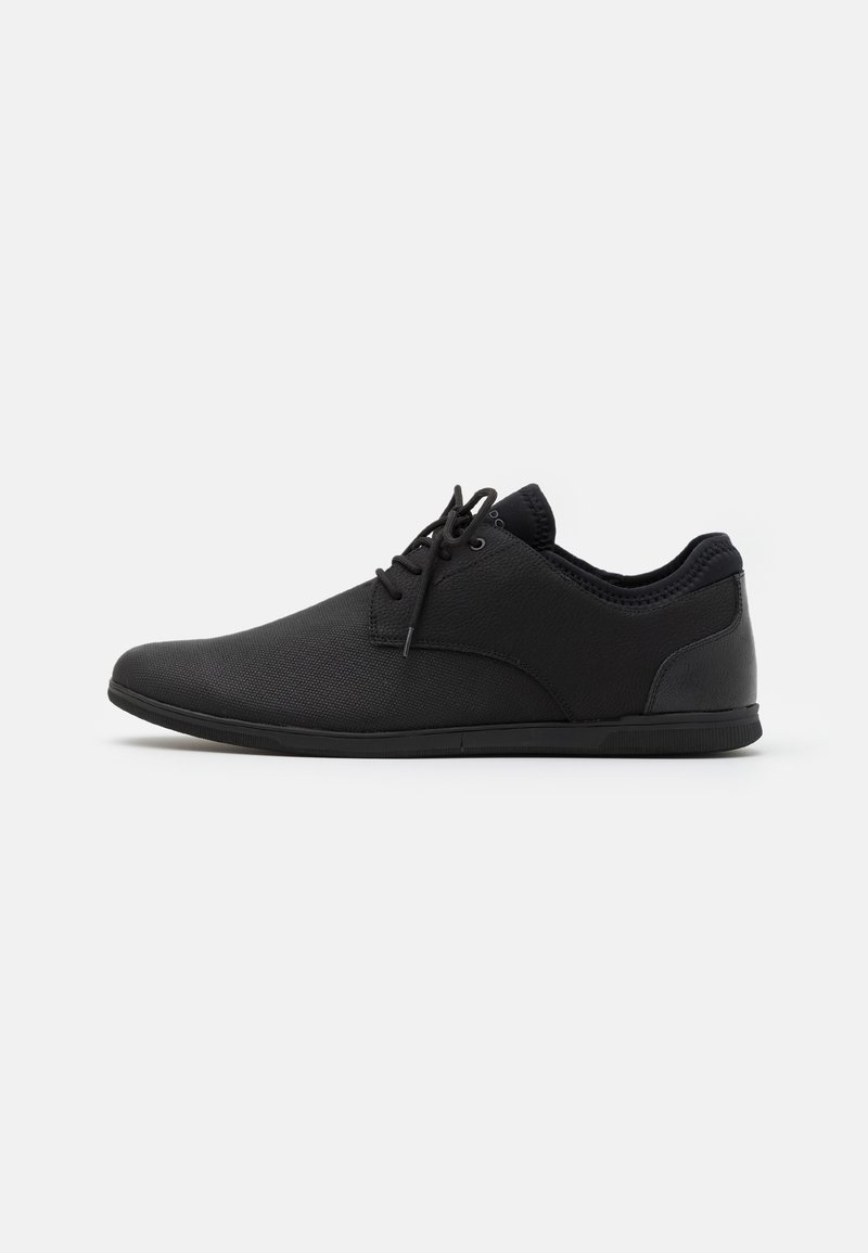 Chaussure décontractée noire basse avec lacets à l'avant et dessus en tissu texturé, photographiée de profil sur un fond clair uni.