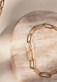 Un collier en chaîne en or repose sur une surface lisse en marbre beige clair, mettant en valeur des maillons ovales allongés avec une finition polie.
