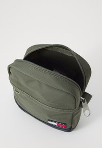 Olivově zelená textilní crossbody taška se zipovým hlavním oddílem, s černým dnem a logem v červené, bílé a modré barvě.