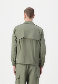 Person with short hair and hoop earrings wearing matching olive green long-sleeve jacket and cargo pants, viewed from behind.