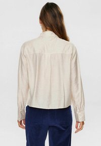 Gai bēšs garām piedurknēm krekls ar teksturētu audumu, pogājamu aproču un īsu apakšu. Iezīmējas ar aizmugurējo jaku un smalkiem daudzkrāsainiem punktiņiem.