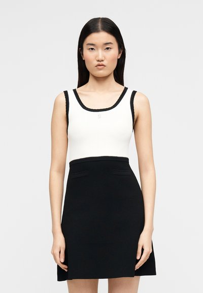 Γυναίκα με ίσια μαύρα μαλλιά που φοράει ένα άσπρο αμάνικο τοπ με μαύρα περιθώρια και μια μαύρη φούστα μέσης ενάντια σε έναν απλό φόντο.