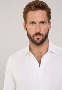 Chemise en lin blanc avec un col boutonné, présentant un tissu texturé et une coupe décontractée, avec des boutons visibles à l'avant.