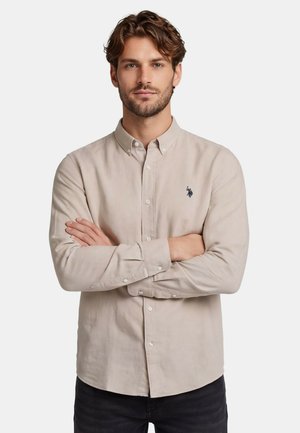 Man with short brown hair and beard wearing a beige button-up shirt with a small dark logo, arms crossed, neutral background.