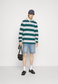 Groen en grijs gestreepte lange mouw polo, blauwe denim shorts, zwarte schoudertas, witte sokken en zwarte instappers, met een grijze pet.