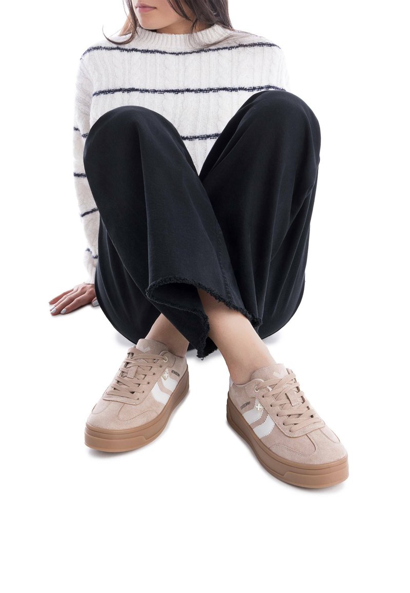 Sneaker scamosciati beige con strisce bianche, punta rotonda e suola in gomma marrone spessa, abbinati a pantaloni neri a gamba larga e a un maglione a righe.