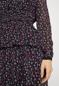 Robe en tissu noir avec petit motif floral rose et blanc, manches longues transparentes, taille cintrée et jupe à volants, main reposant sur le côté.