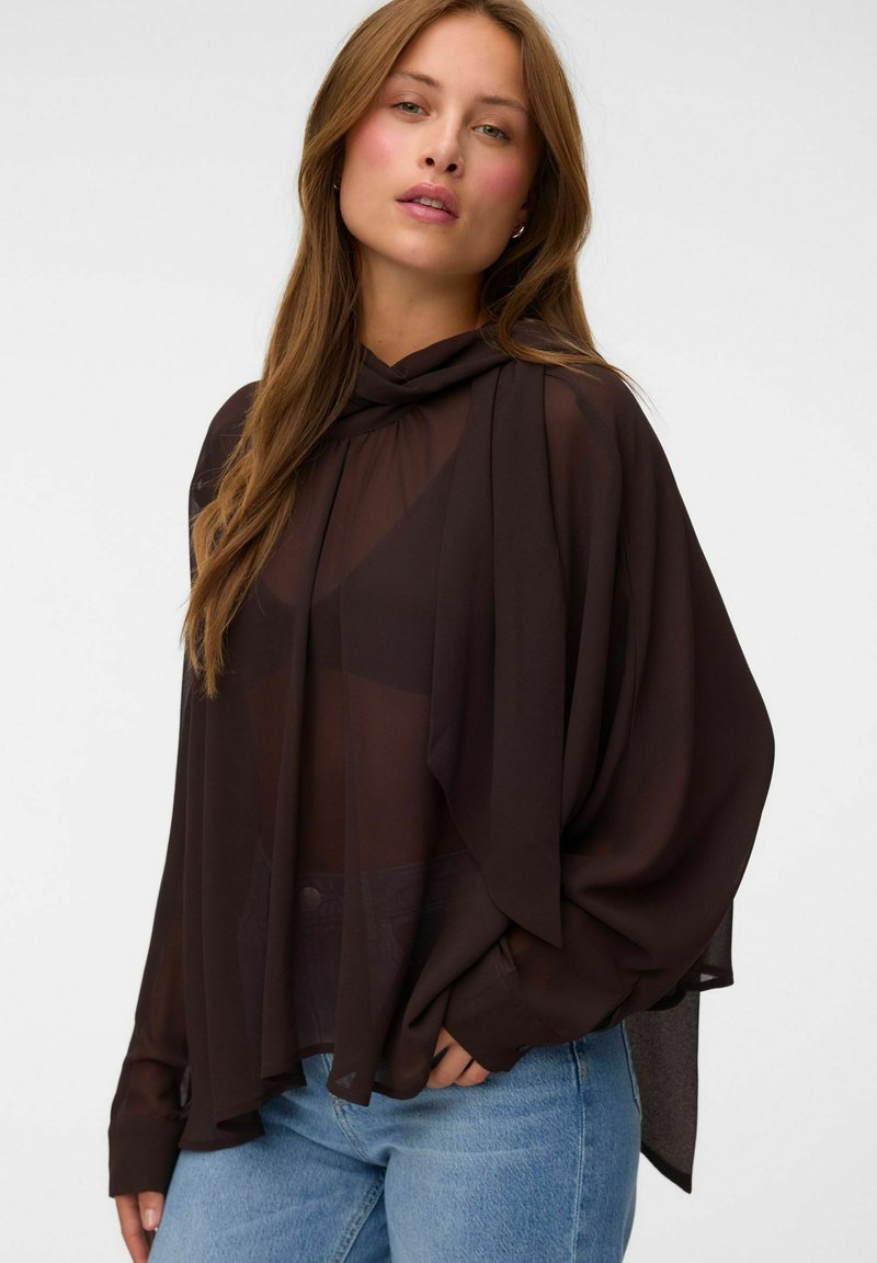 Femme aux longs cheveux châtain clair porte un chemisier noir transparent sur un top noir et un jean bleu, debout devant un fond clair uni.