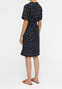 Robe bleu marine à pois avec des manches courtes, taille à nouer, coupe genou et texture lisse. Portée avec des sandales à enfiler orange.