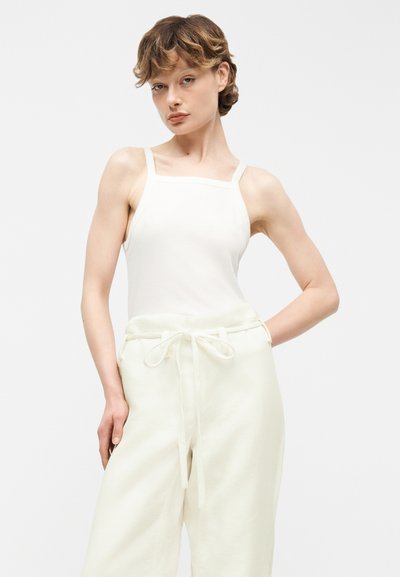 Femme aux cheveux courts et bouclés portant un haut blanc sans manches à col carré et un pantalon blanc taille haute noué avec un cordon, posant.