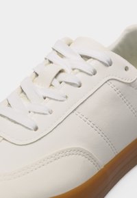 Λευκό δερμάτινο αθλητικό παπούτσι με λευκά κορδόνια και ελαφρώς καφέ καουτσούκ σόλα, με γωνία που δείχνει τις ραφές στο πλάι και στο πάνω μέρος.