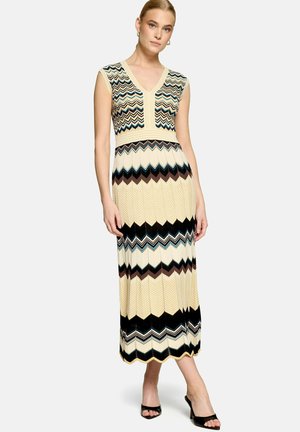 Femme portant une robe longue sans manches à col en V avec des motifs zigzag multicolores en crème, noir, marron et bleu, associée à des talons ouverts noirs.