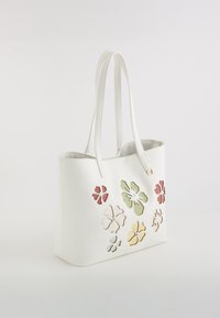 Witte leren tote bag met bloemen uitgesneden ontwerpen in roze, groen, geel en blauw; dubbele schouderbanden; gladde textuur.