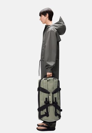 Giacca impermeabile grigia con cappuccio, borsa da trekking verde con cinghie nere, tenuta da una persona con infradito, posizionata di lato.