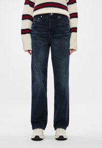 Jeans din denim albastru închis, cu o croială relaxată, design cu cinci buzunare și decolorare subtilă. Purtați cu un pulover crem și adidași.