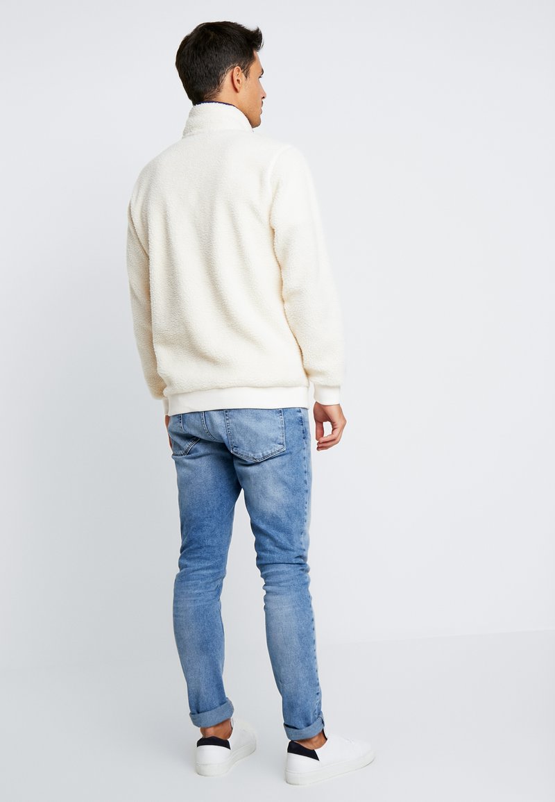 Witte fleece pullover met een hoge kraag, gedragen met lichtblauwe jeans; beschikt over geribde manchetten en zoom, en heeft een relaxte pasvorm.