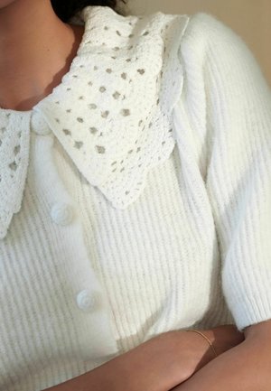 Bílý pletený cardigan s ribbovanou texturou, s velkým límcem ozdobeným krajkou a třemi knoflíkovými uzávěry vpředu.