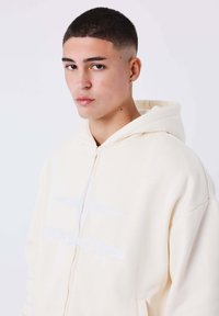 Sudadera de color crema con una textura suave, que presenta un detalle de logo bordado en blanco. La capucha es amplia y ligeramente sobredimensionada.
