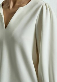 Lichtbeige blouse met een V-hals en pofmouwen, gemaakt van een gladde stof met een zachte textuur en subtiele glans. Geen opvallende patronen of hardware.