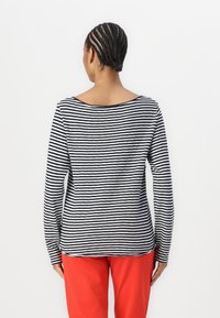 Langärmliges gestreiftes Hemd in Marineblau und Weiß, mit weitem Ausschnitt und lässigem Schnitt. Das Material wirkt weich; der Saum ist leicht abgerundet. Kombiniert mit roten Hosen.