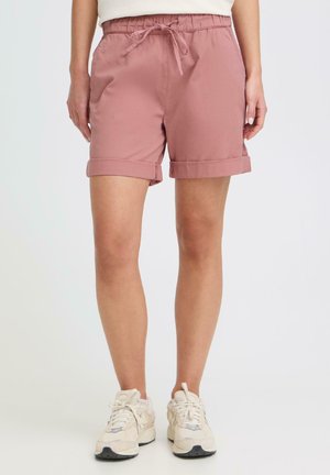 Femme portant un short rose poudré à cordon de serrage avec ourlets retroussés et des baskets blanches, debout devant un fond uni clair.